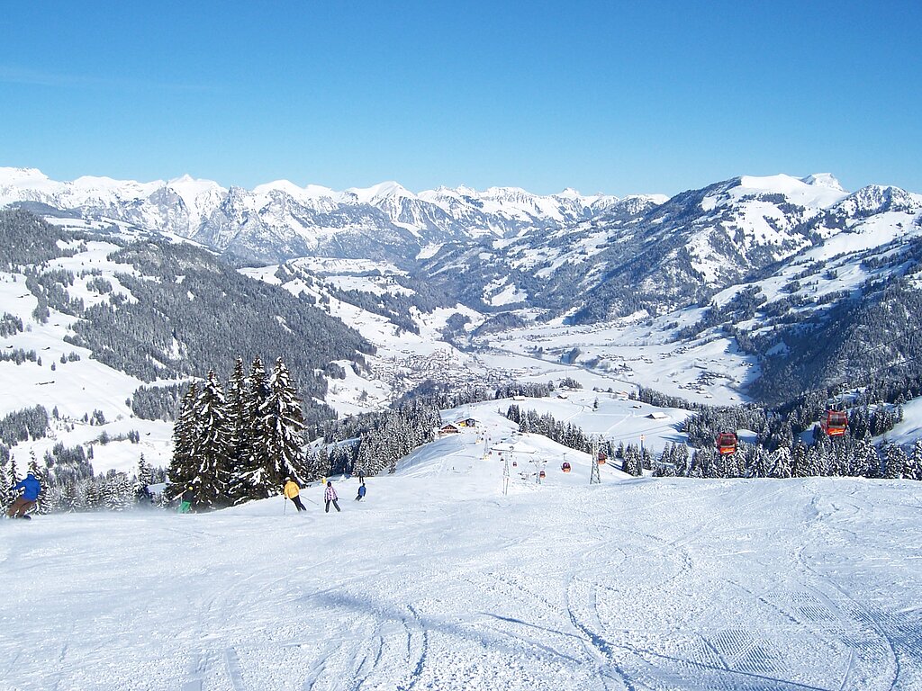 Skipiste von oben Richtung Tal fotografiert, im Hintergrund liegt Zweisimmen umgeben von verschneiten Bergen.