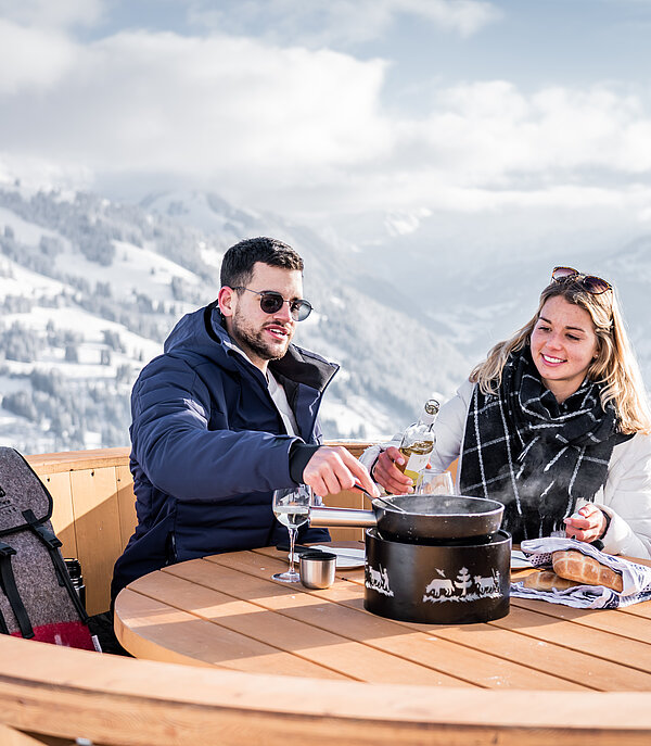 Ein Paar sitzt in einem grossen Fondue Caquelon aus Holz und geniesst ein Fondue aus dem Fonduerucksack sowie die verschneite Berglandschaft im Hintergrund.