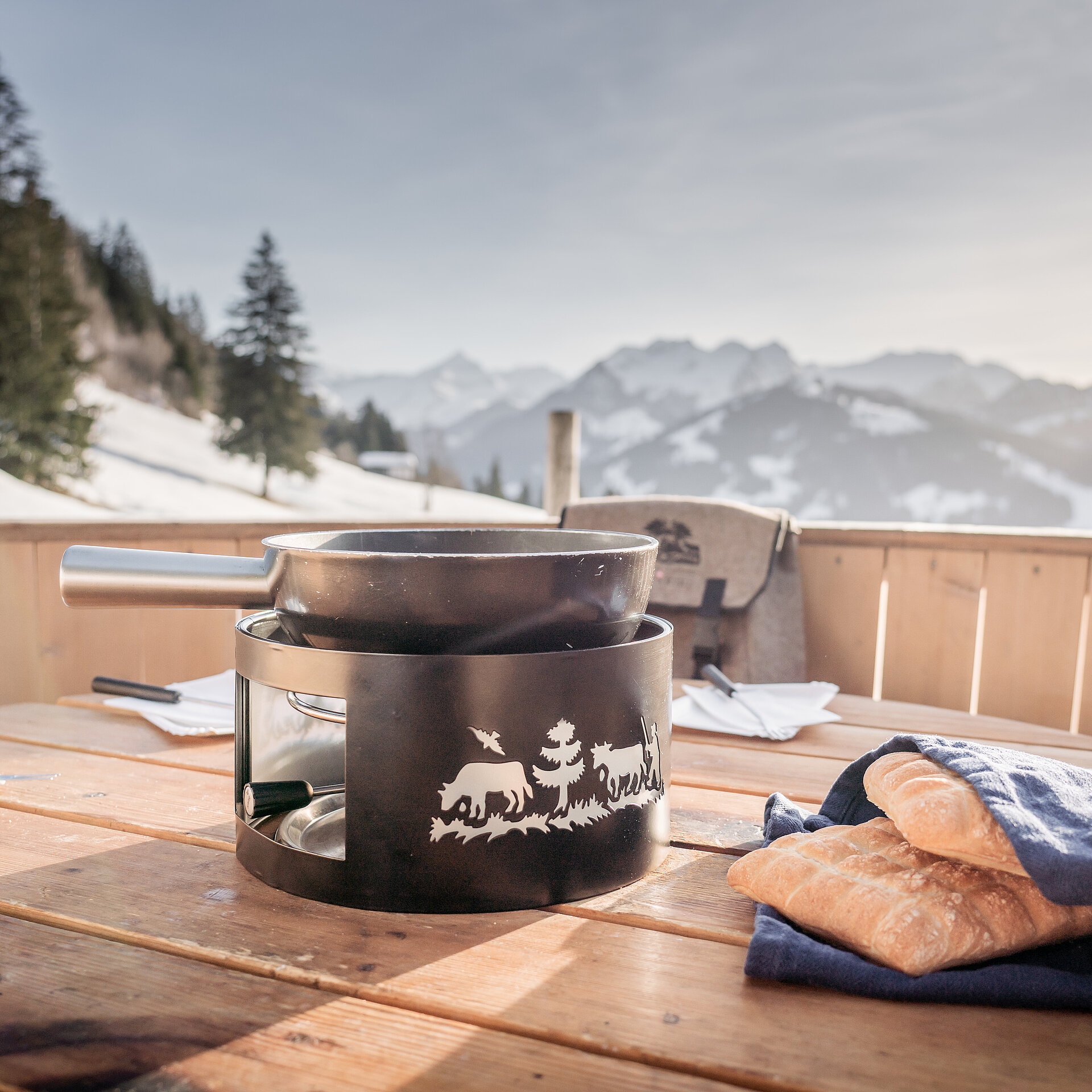 Schwarzes Fonduecaquelon und Brot stehen im überdimensionalen Fonduecaquelon aus Holz bereit, Aussicht auf winterliche Landschaft mit Bäumen und Bergen.