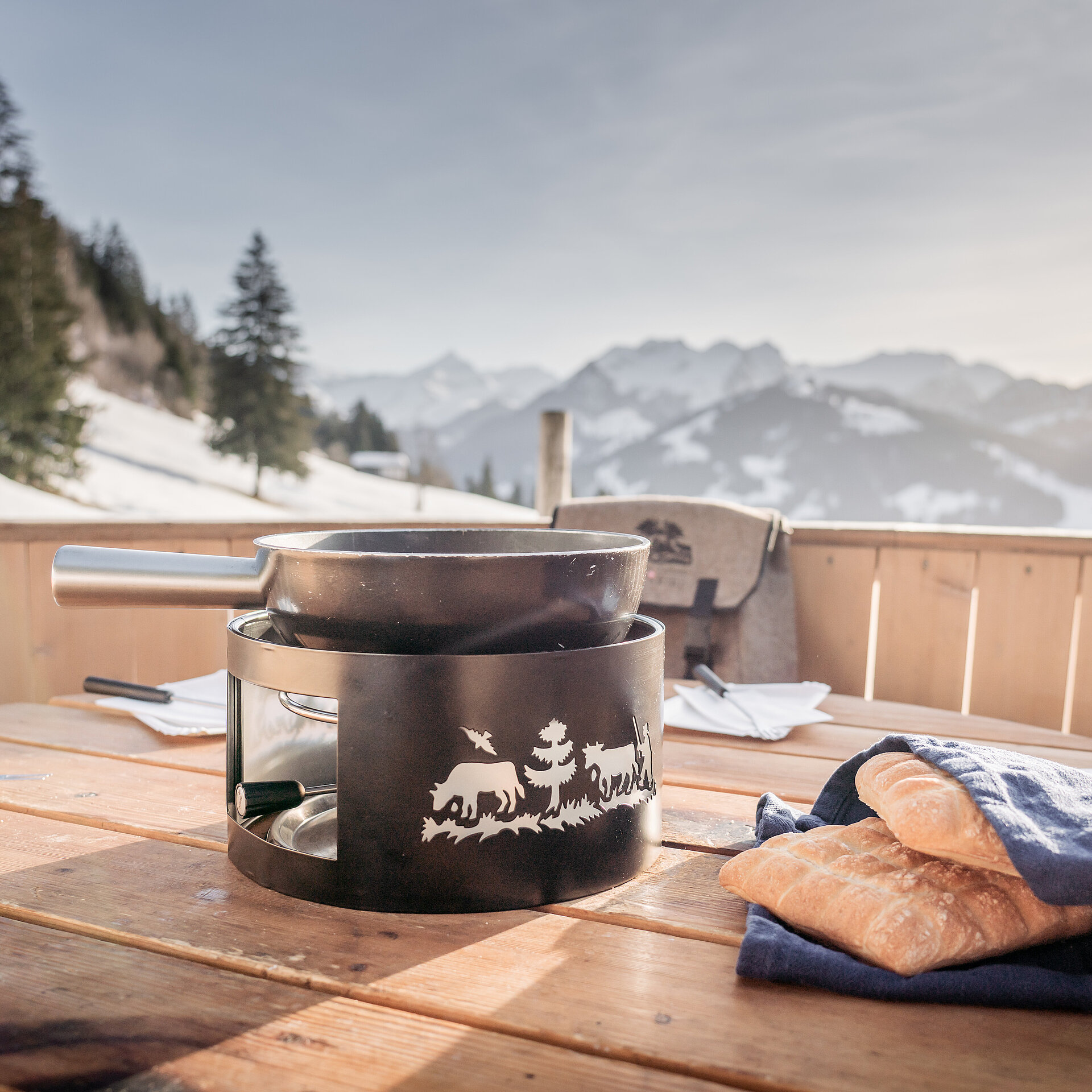 Schwarzes Fonduecaquelon und Brot stehen im überdimensionalen Fonduecaquelon aus Holz bereit, Aussicht auf winterliche Landschaft mit Bäumen und Bergen.