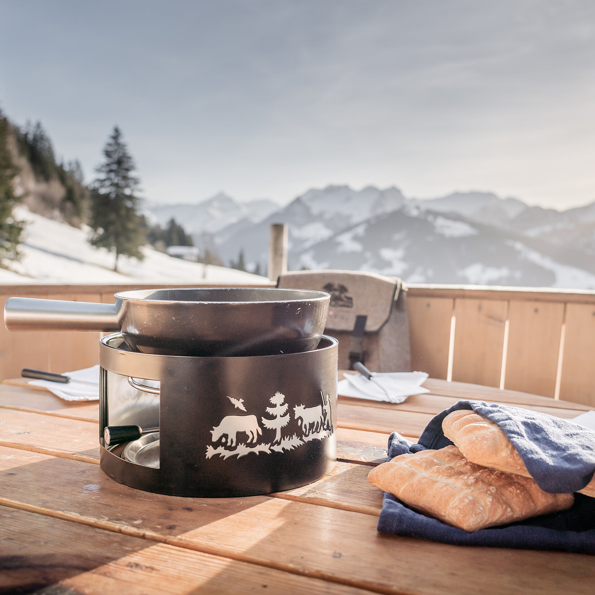 Schwarzes Fonduecaquelon und Brot stehen im überdimensionalen Fonduecaquelon aus Holz bereit, Aussicht auf winterliche Landschaft mit Bäumen und Bergen.