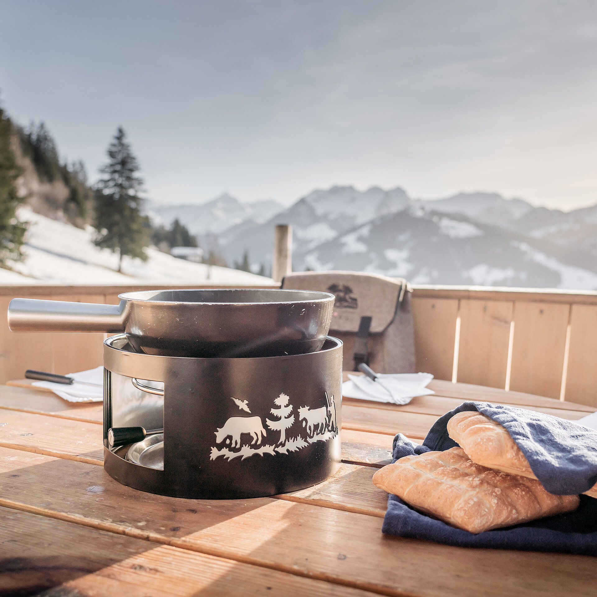 Schwarzes Fonduecaquelon und Brot stehen im überdimensionalen Fonduecaquelon aus Holz bereit, Aussicht auf winterliche Landschaft mit Bäumen und Bergen.