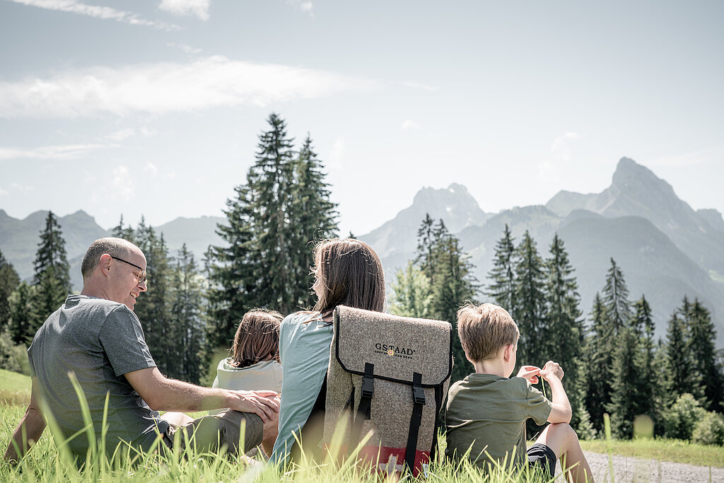 Eine Familie mit Mann, Frau, Tochter und Sohn sitzen in einer grünen Wiese an einem Wegrand im Hintergrund Bergpanorama. Die Frau trägt einen Fonduerucksack.