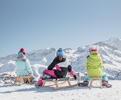 Drei Kinder sitzen auf hölzernen Schlitten und bewundern die verschneiten Berge im Hintergrund.