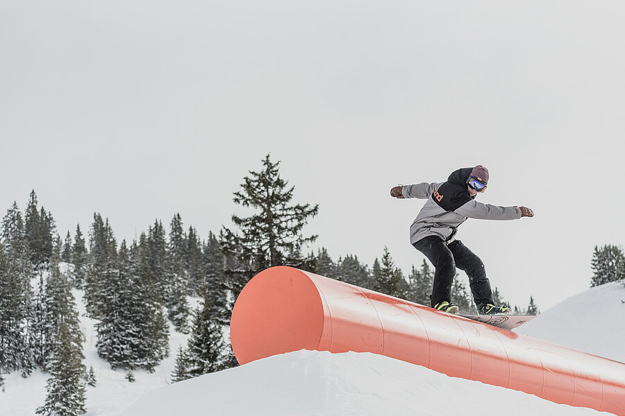 Ein dunkel gekleideter Snowboarder fährt über ein orangenes Rail im Snowpark.