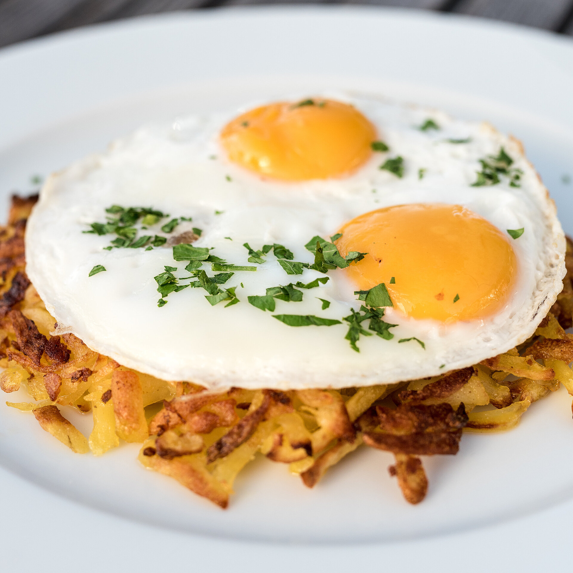 Weisser Teller mit braun gebrannter Rösti und zwei Spiegeleiern garniert mit Kräutern.