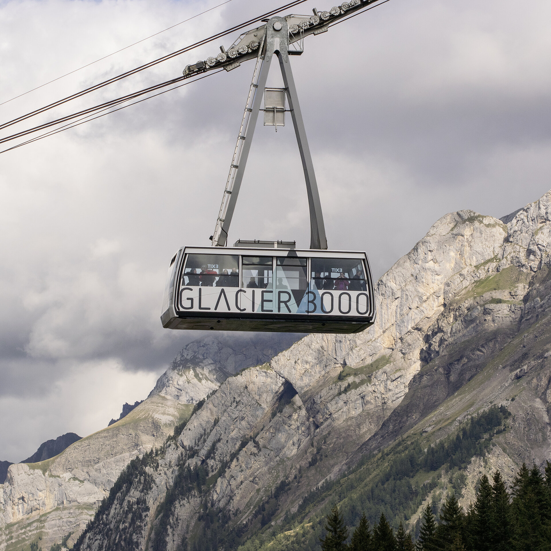 Luftseilbahn mit Aufschrift Glacier 3000 am Seil vor sommerlicher Bergkulisse und bewölktem Himmel.