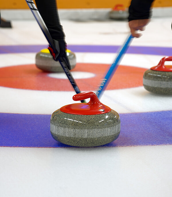 Zwei Curlingspieler wischen einen rot-grauen Curlingstein in den blau-weiss-roten Ring. Daneben liegen drei weitere Curlingsteine.