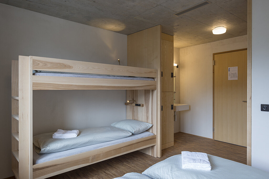 Famililenzimmer mit einem Etagenbett und im Vordergrund ein Teil eines Doppelbettes mit weisser Bettwäsche. Im Hintergrund ein Schrank, ein Lavabo und eine Türe