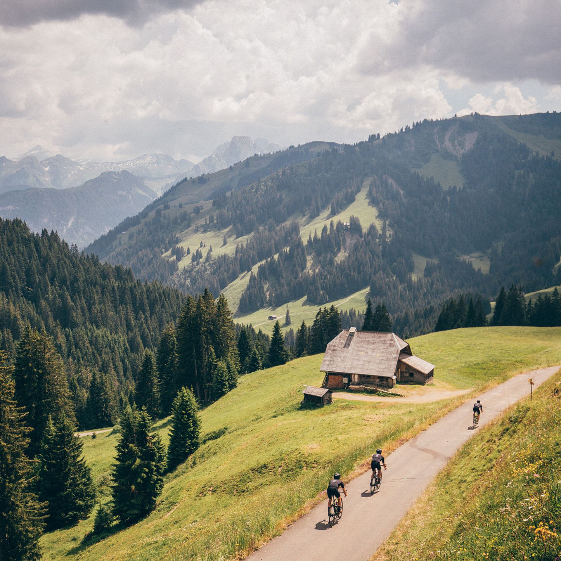 Drei Radsportler fahren vom Mittelbergpass auf der schmalen Strasse ins Grischbachtal. Im Hintergrund sieht man Bergpanorama und viele Wolken.