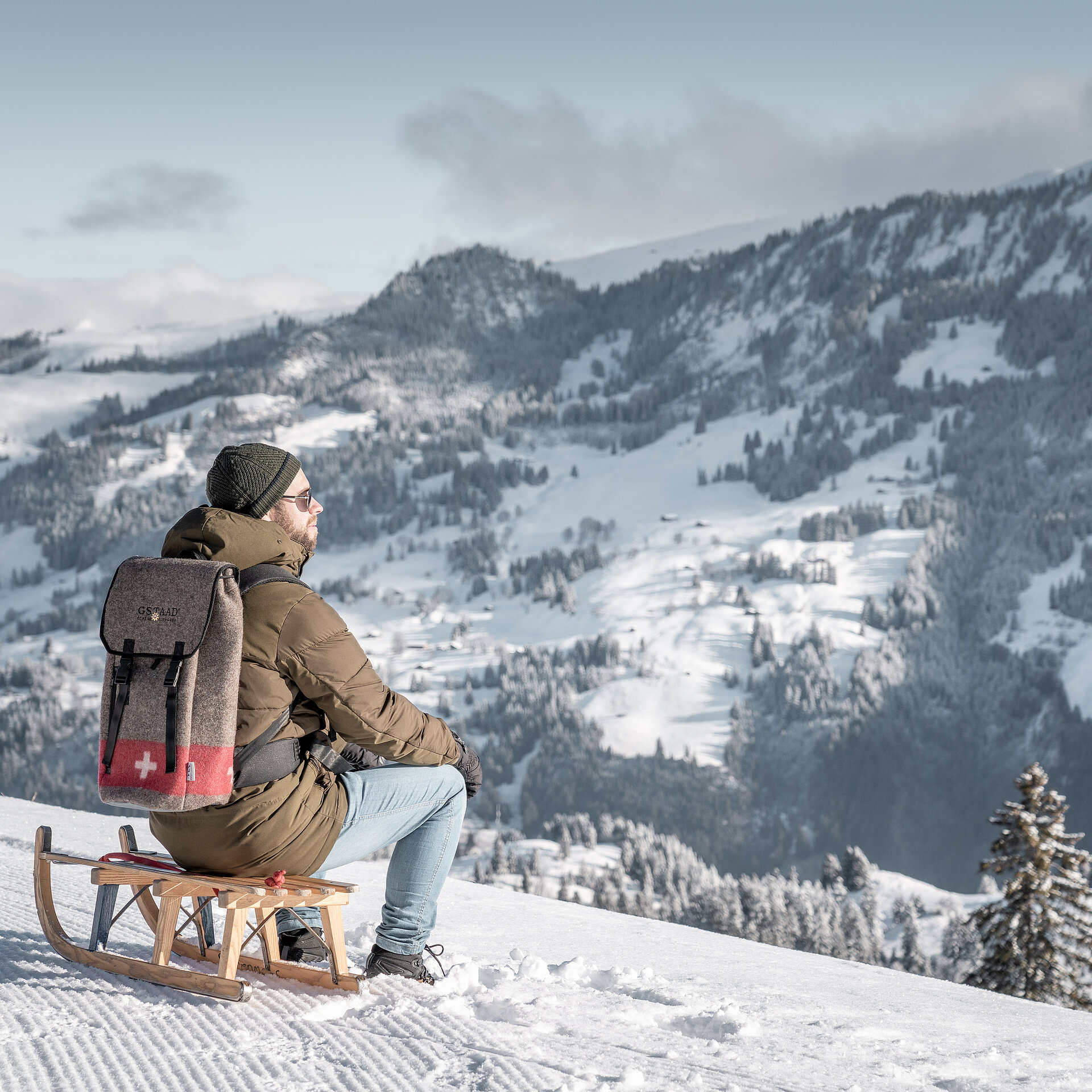 Person sitzend auf einem Holzschlitten vor verschneiter Bergkulisse.