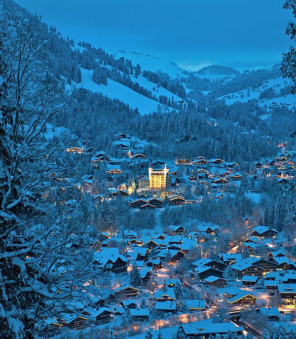 Blick auf das verschneite Dorf Gstaad mit dem beleuchteten Gstaad Palace und Berge im Hintergrund.