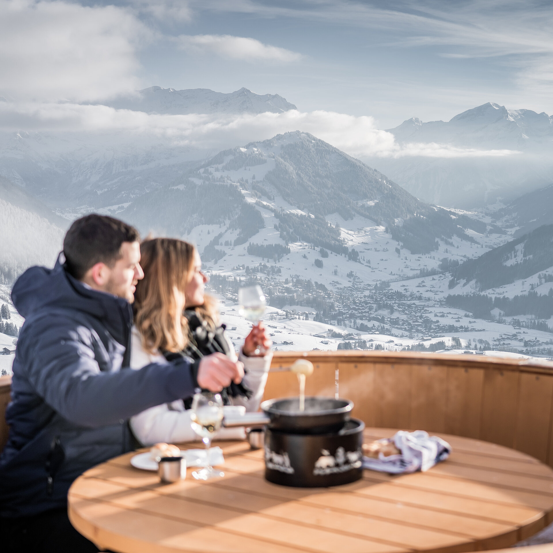 Ein Paar sitzt in einem grossen Fondue Caquelon aus Holz, geniesst ein Fondue und die Aussicht auf die Berge.