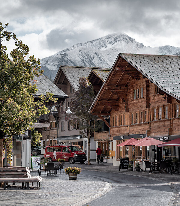 Dorfzentrum von Saanen mit typischen alten Chalets, Sitzbänken und einem Baum im Vordergrund und verschneiten Bergen im Hintergrund.