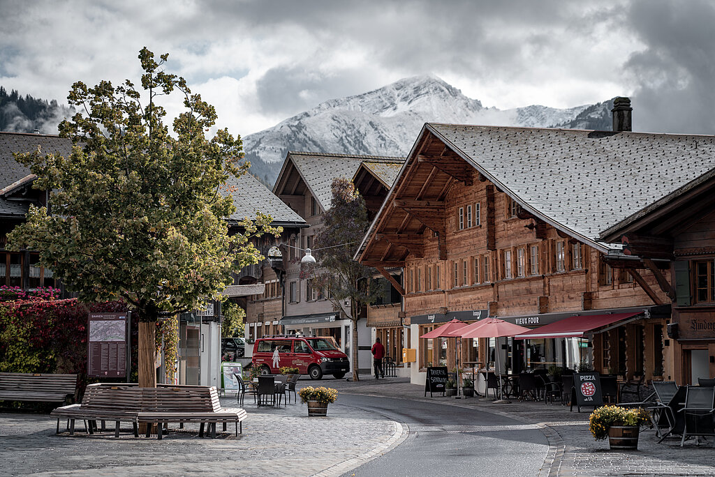Dorfzentrum von Saanen mit typischen alten Chalets, Sitzbänken und einem Baum im Vordergrund und verschneiten Bergen im Hintergrund.