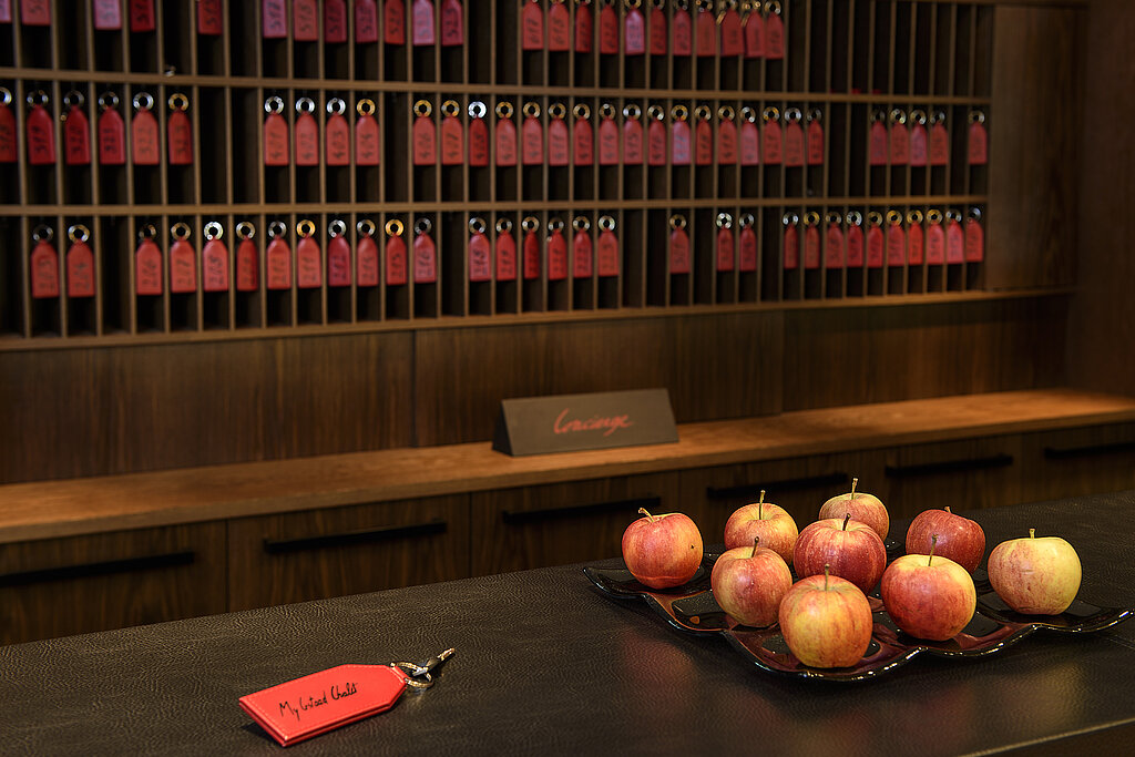 Empfangstheke mit einem Zimmerschlüssel mit rotem Anhänger und einer Schale mit Äpfeln, im Hintergrund Schlüsselbrett mit Zimmerschlüsseln.