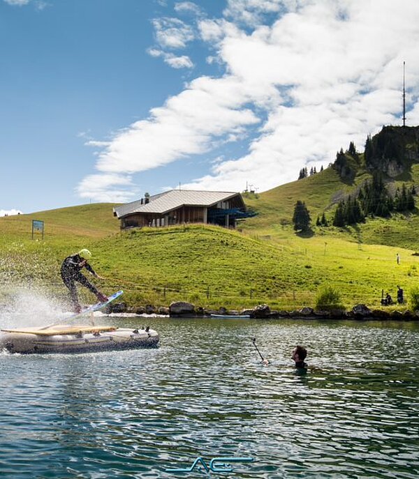 Eine Person auf einem Wakeboard fährt auf einem kleinen Bergsee über verschiedene Schanzen.