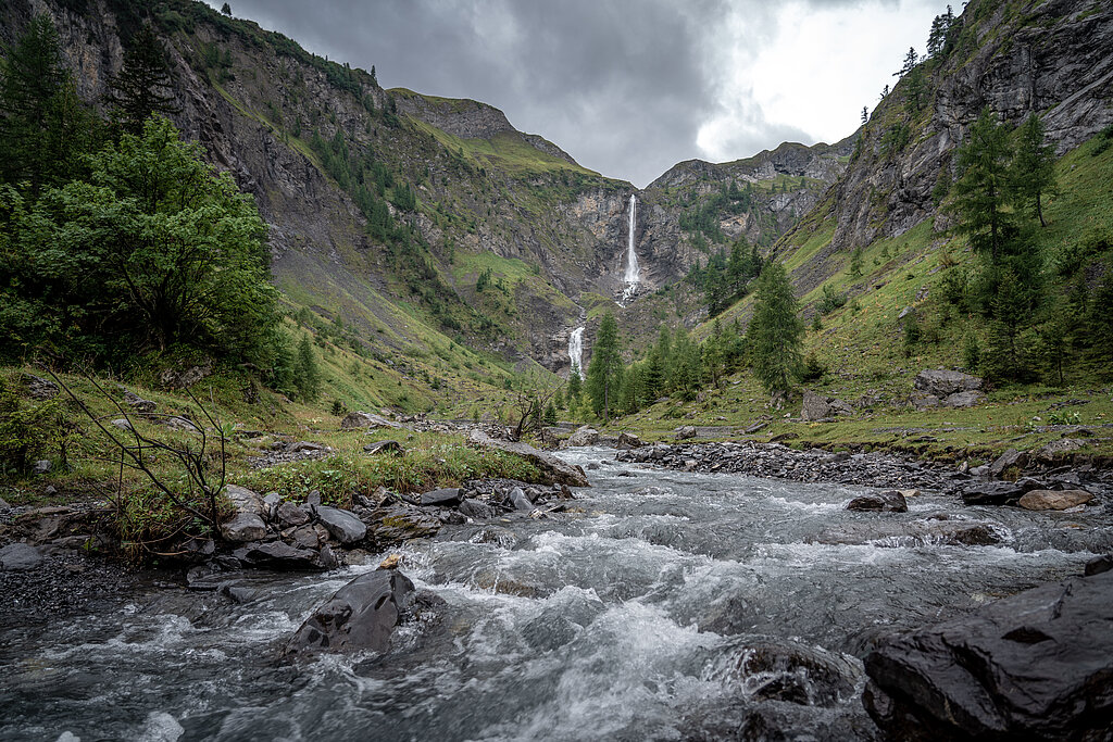 Ein imposanter Wasserfall stürzt sich ins Tal hinunter. Im Vordergrund ein Bach, dahinter Berge.