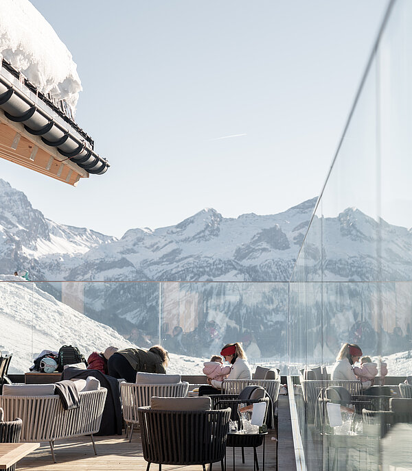 Gäste sitzen im Winter auf Bergrestaurantterasse, welche mit Glaswänden vor Wind schützt. Schöne weisse Berglandschaft im Hintergrund.