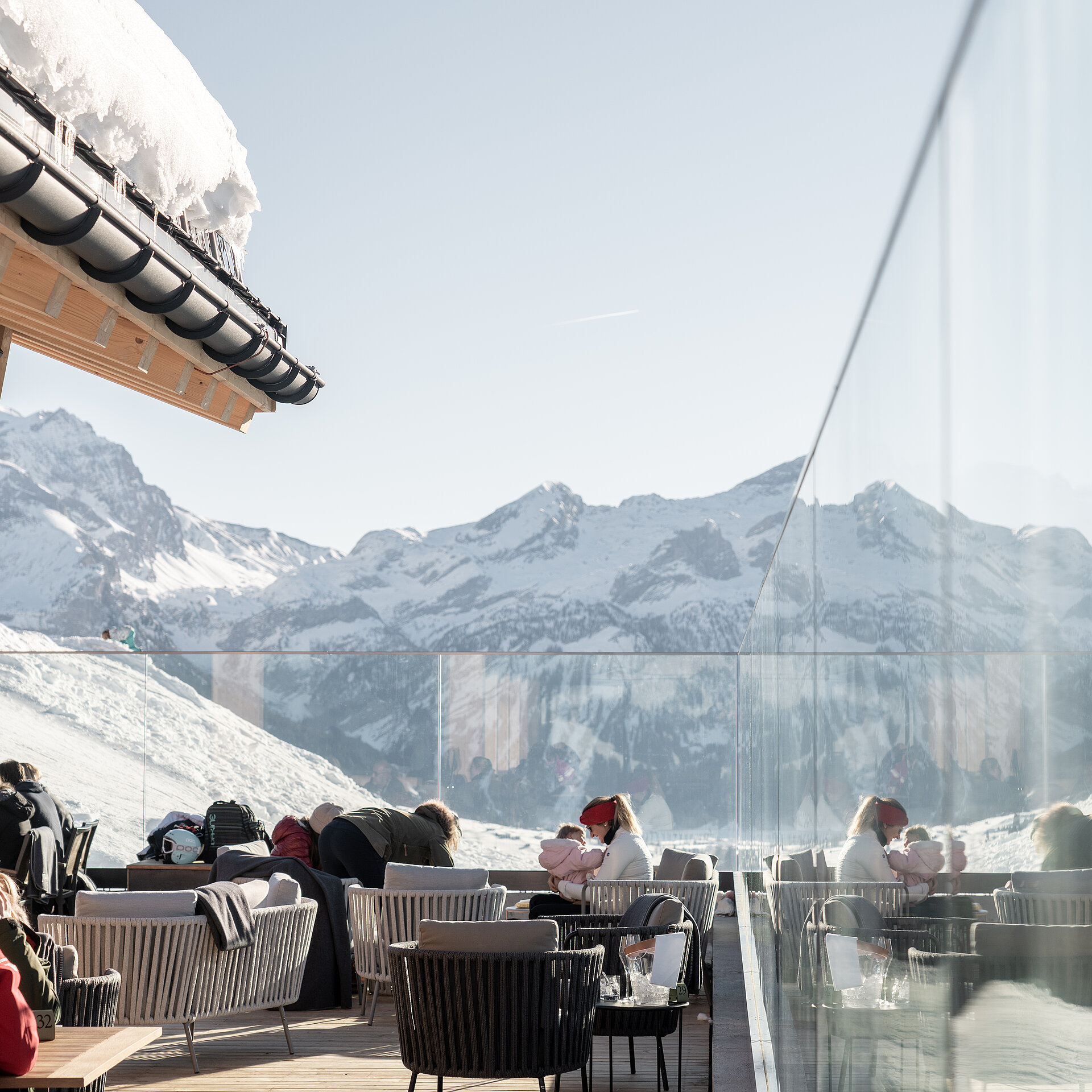 Gäste sitzen im Winter auf Bergrestaurantterasse, welche mit Glaswänden vor Wind schützt. Schöne weisse Berglandschaft im Hintergrund.