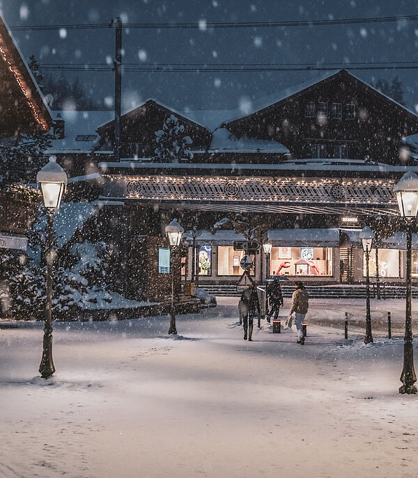 Drei Menschen laufen am Abend durch die romantisch beleuchtete Promenade in Gstaad, während der Schnee fällt.