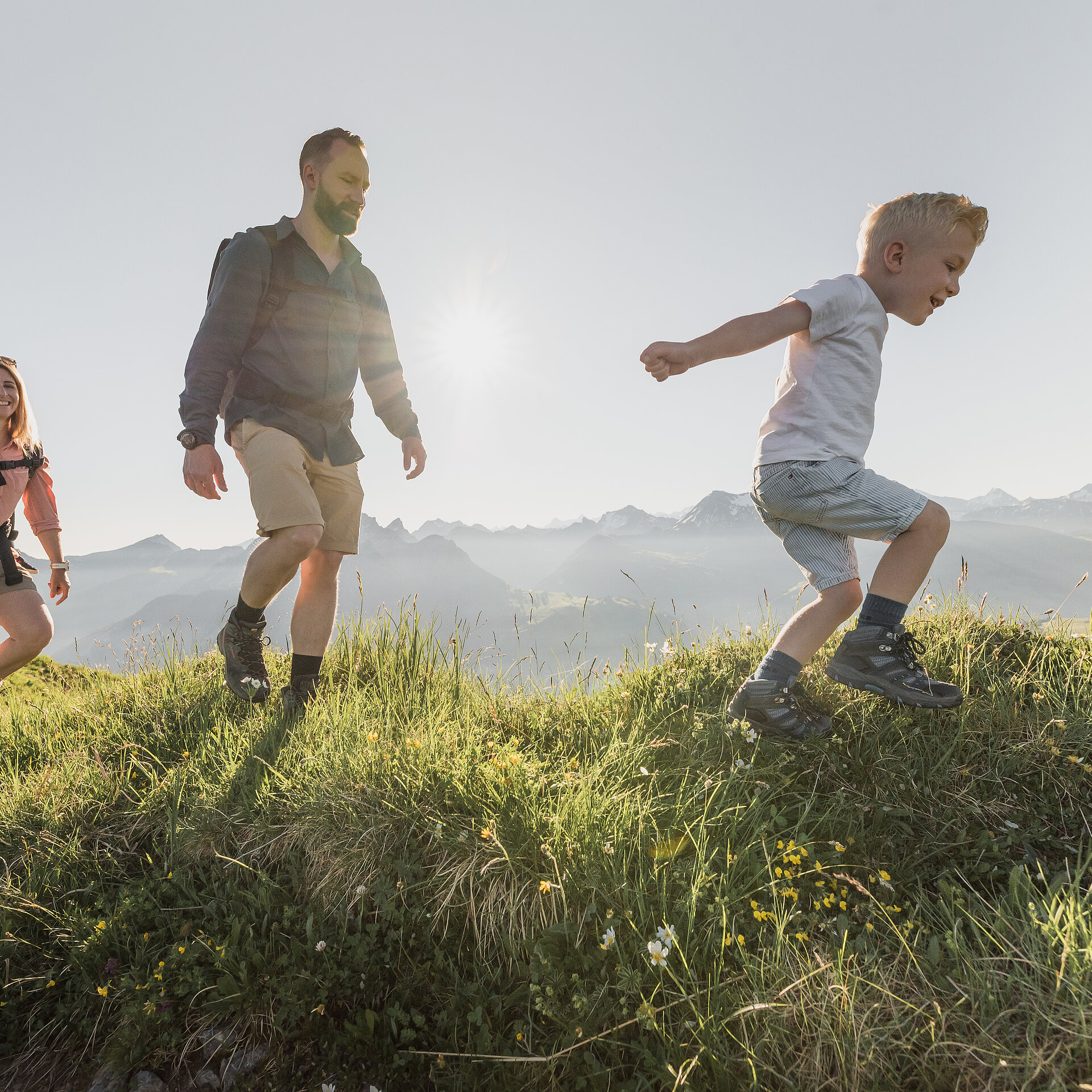 Familie bestehend aus drei Personen beim Wandern über grüne Wiesen bei sommerlichem Wetter.