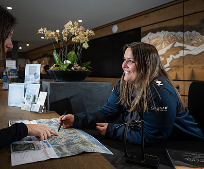 Eine Mitarbeiterin von Gstaad Saanenland Tourismus zeigt einem weiblichen Gast etwas auf einer Panoramakarte. Im Hintergrund die Schalter des Tourismusbüros Gstaad.