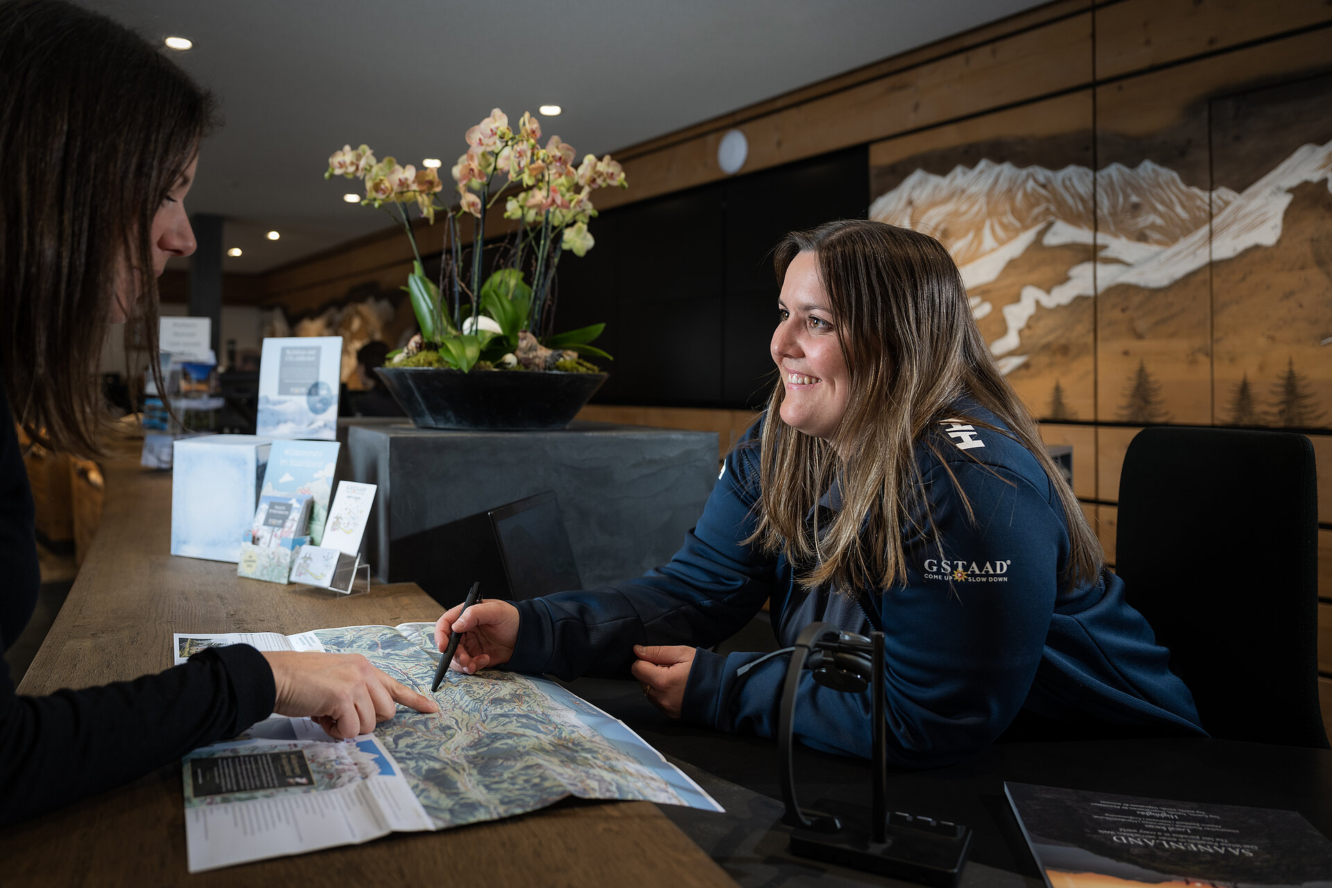 Eine Mitarbeiterin von Gstaad Saanenland Tourismus zeigt einem weiblichen Gast etwas auf einer Panoramakarte. Im Hintergrund die Schalter des Tourismusbüros Gstaad.