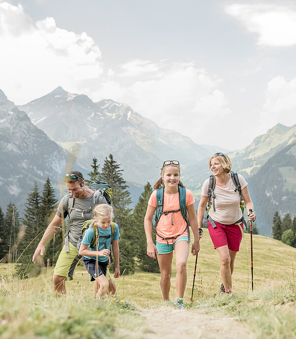 Eine Familie wandert auf einem Wanderweg neben einer grünen Wiese, im Hintergrund eine Berglandschaft.