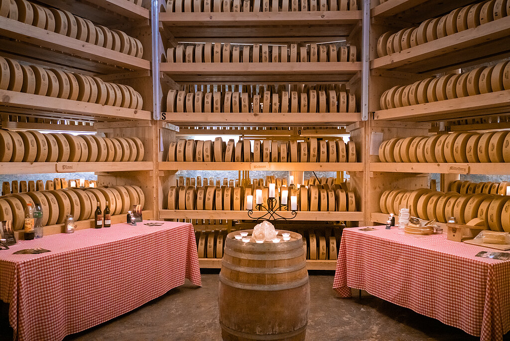 Hintergrund mit Regalen voller Käse, in der Mitte stehen Tische bereit für die Degustation.