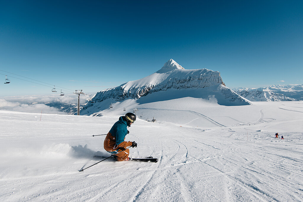 Ein Skifahrer in orange-türkis farbigem Skianzug fährt auf einer Skipiste entlang eines Sesselliftes mit Blick auf den Gletscher und das Bergpanorama.