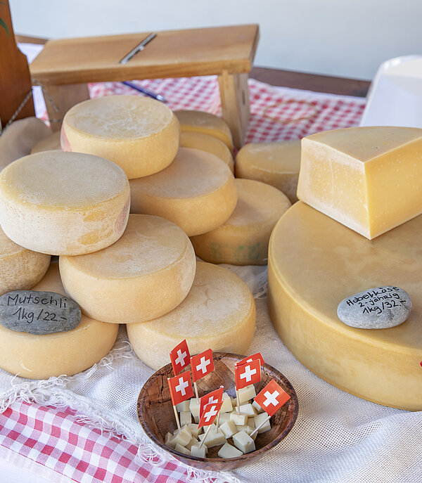 Auf einem Tisch mit rot/weissem Tischtuch liegen verschiedene Käsesorten. Zehn kleine Mutschli, ein grosser Käse mit einem Abschnitt obendrauf und in einem Korb ist Hobelkäse. Auf dem Tisch ist auch noch ein Hobel und einen kleinen Blumenstrauss zu sehen.