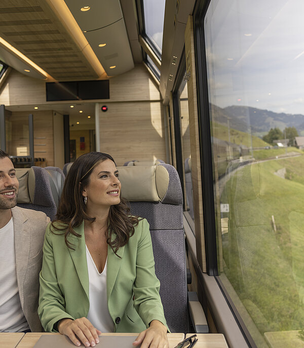 Ein Mann und eine Frau sitzen in einem Zugabteil und schauen aus dem grossen Panoramafenster in die grüne Landschaft