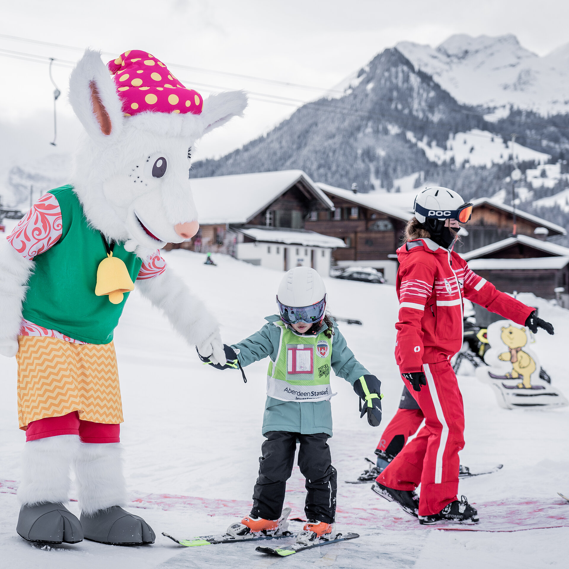 Das Maskottchen Saani im Kinderland der Skischule Gstaad mit einer rot gekleideten Skilehrerin und zwei kleinen Kindern auf Ski.