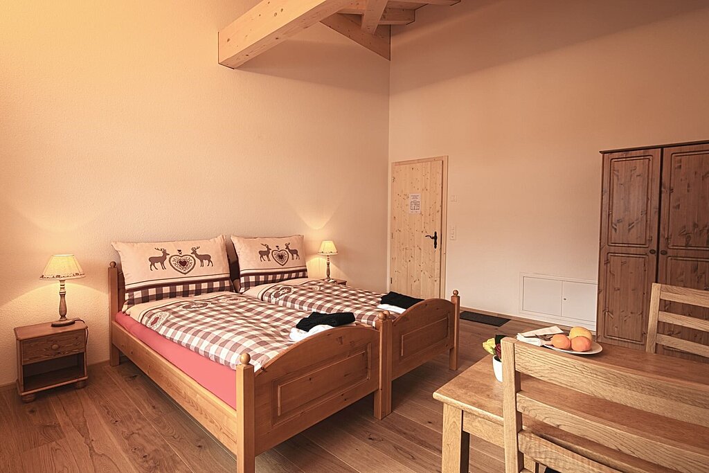 Holziges Doppelbett mit Bettwäsche im Landhausstil und zwei Nachttischen mit Lampe. Rechts ein Tisch und zwei Stühle. Im Hintergrund eine helle Holztüre und ein dunkelbrauner Holzschrank.