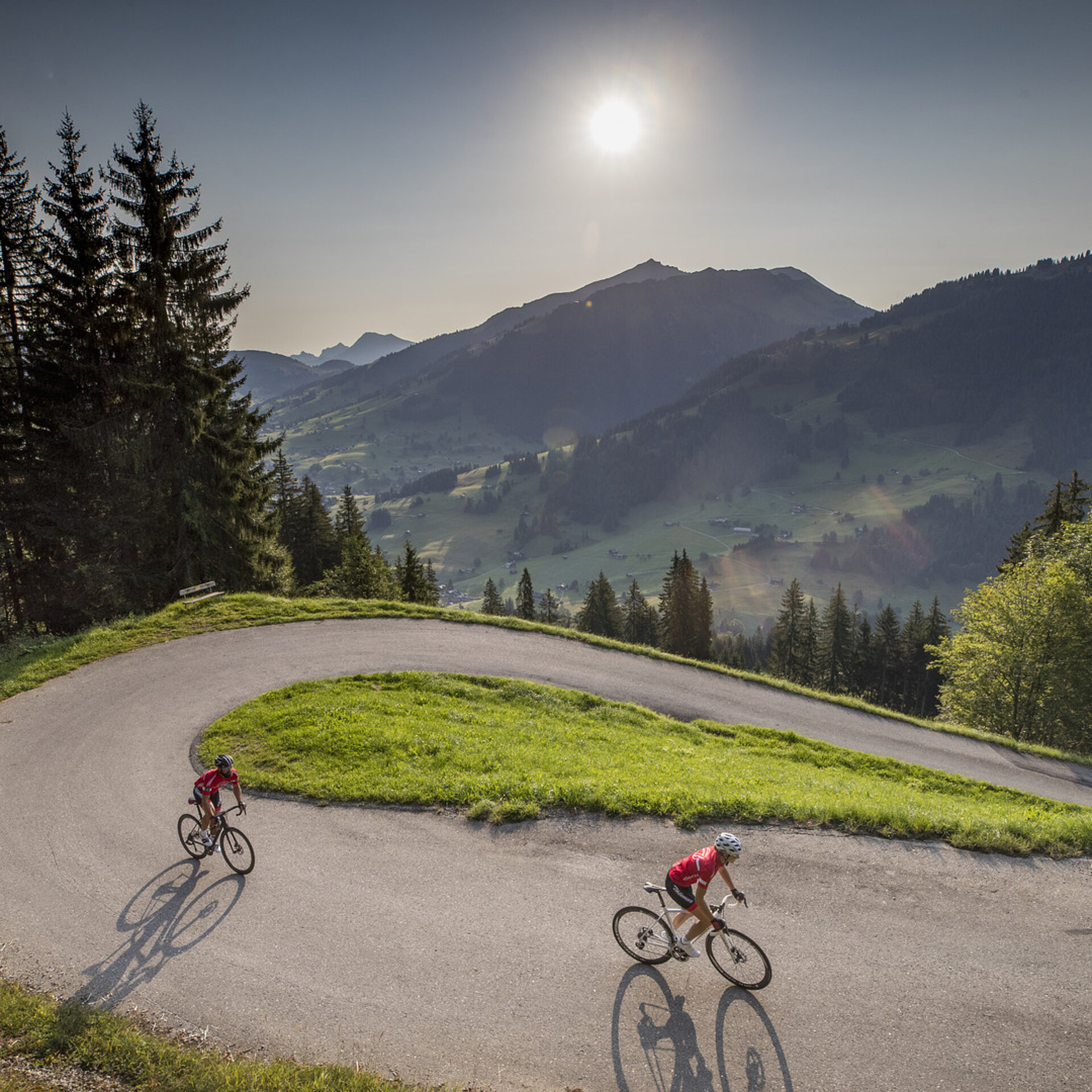 Zwei Rennradfahrer (eine Frau und eine Mann) fahren in einer steilen Kehre bergauf. Am Himmel sieht man die Sonne in einem milchig-blauen Himmel. Es ist Sommer.