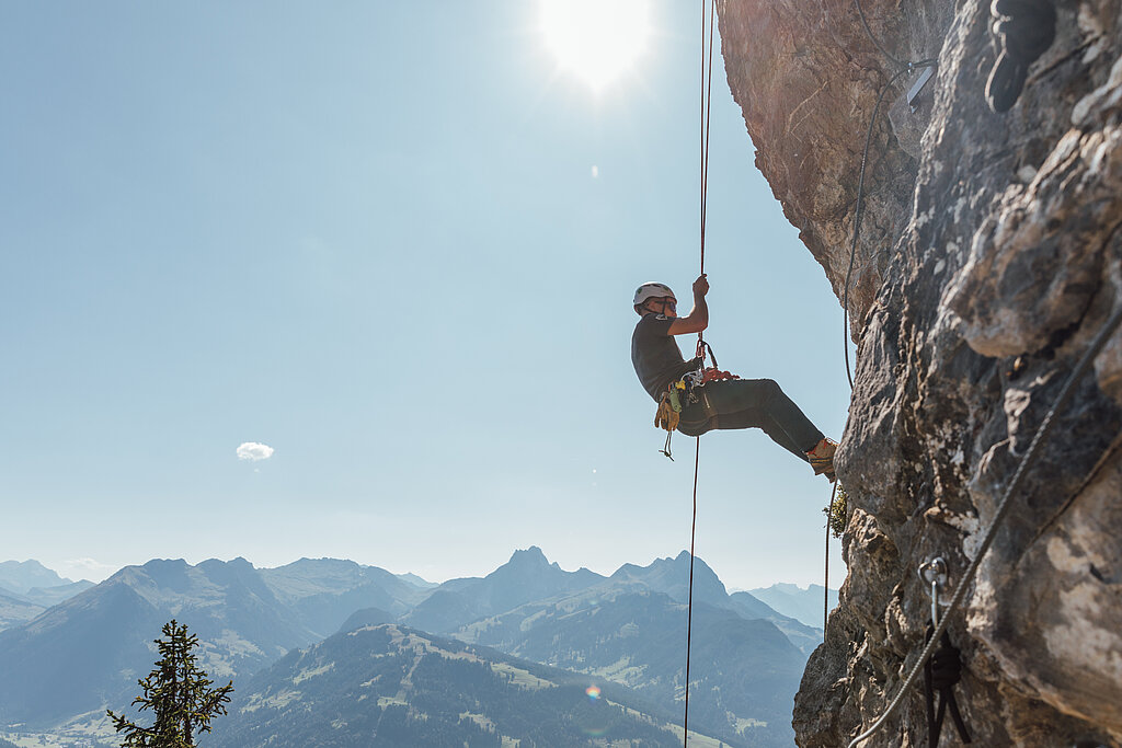 Ein Kletterer seilt sich an einem Seil den Fels hinab, im Hintergrund die Bergkulisse vom Saanenland unter blauem Himmel.