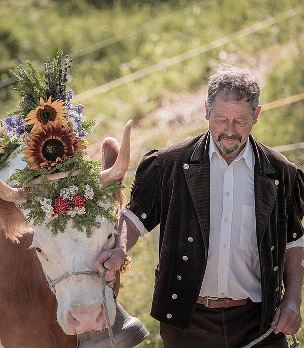 Ein Bauer in traditioneller Alpkleidung mit schwarzer Jacke und weissem Hemd führt eine Kuh an der Hand. Die Kuh ist mit wunderschönen Blumen geschmückt.