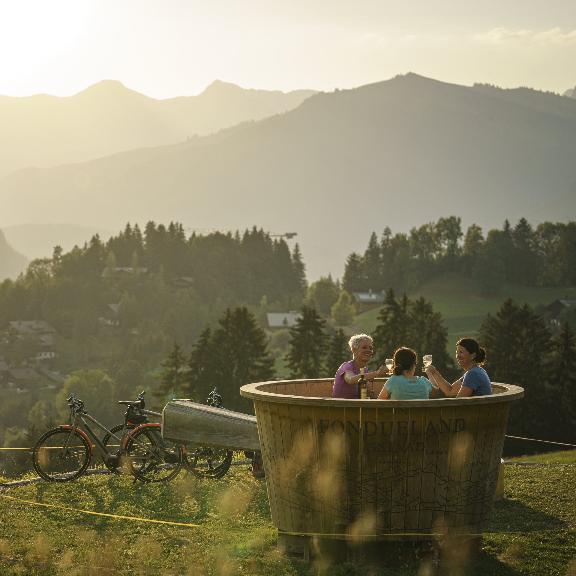 Inmitten schöner Berglandschaft sitzen drei Frauen in einem überdimensionalen Fonduecaquelon aus Holz und prosten sich zu. Daneben stehen ihre Fahrräder.