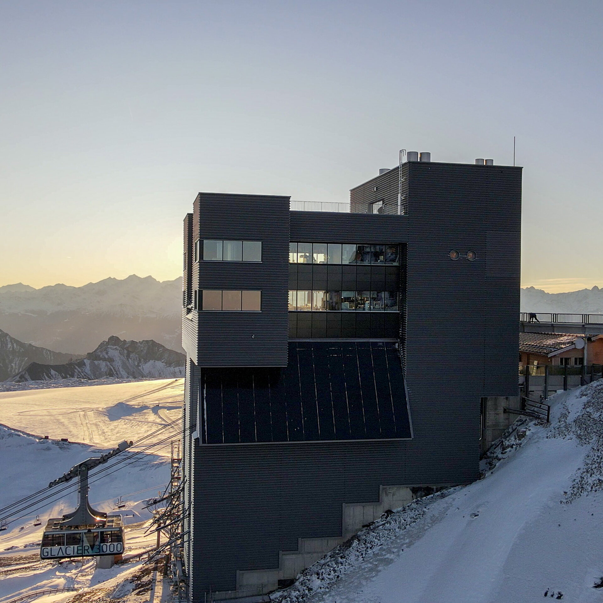 Dunkles hohes Gebäude. Links eine Luftseilbahnkabine und eine Sesselbahn. Rechts ein Holzchalet und eine Brücke. Im Hintergrund verschneite Berge und der Gletscher.