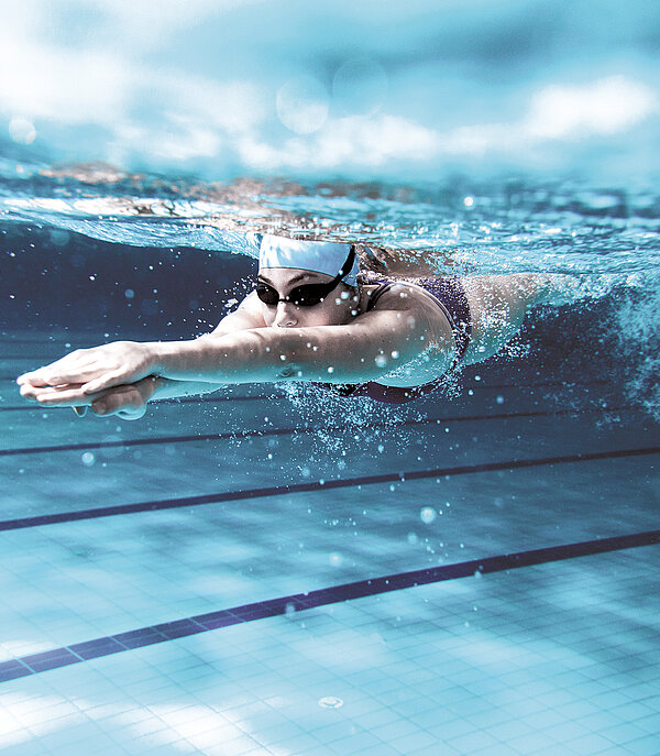 Sportzentrum Gstaad Eine Schwimmerin mit Schwimmmütze und Brille taucht in einem Hallenbad.