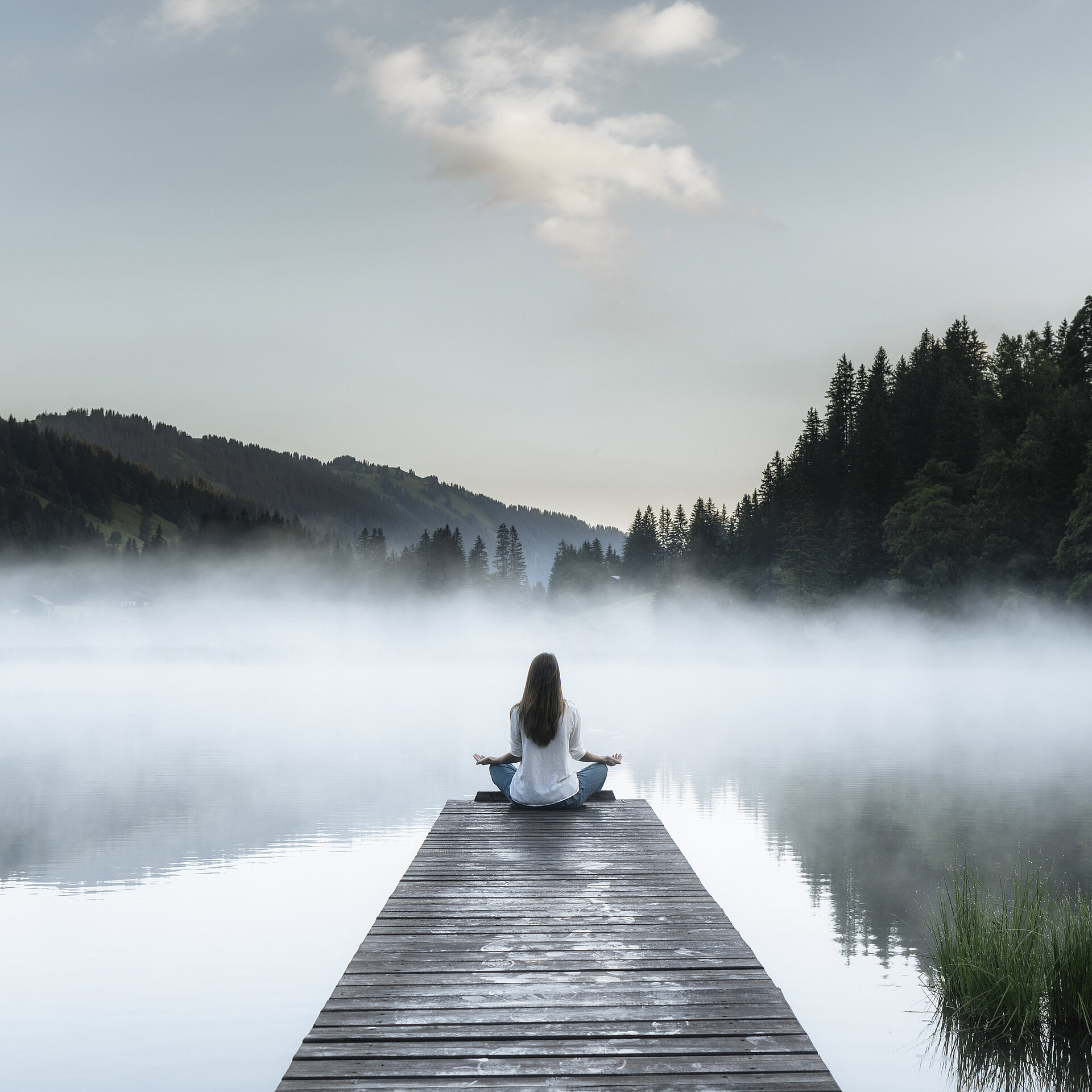 Frau macht Entspannungsübungen auf einem Steg beim See, welcher leicht im Nebel eingehüllt ist.