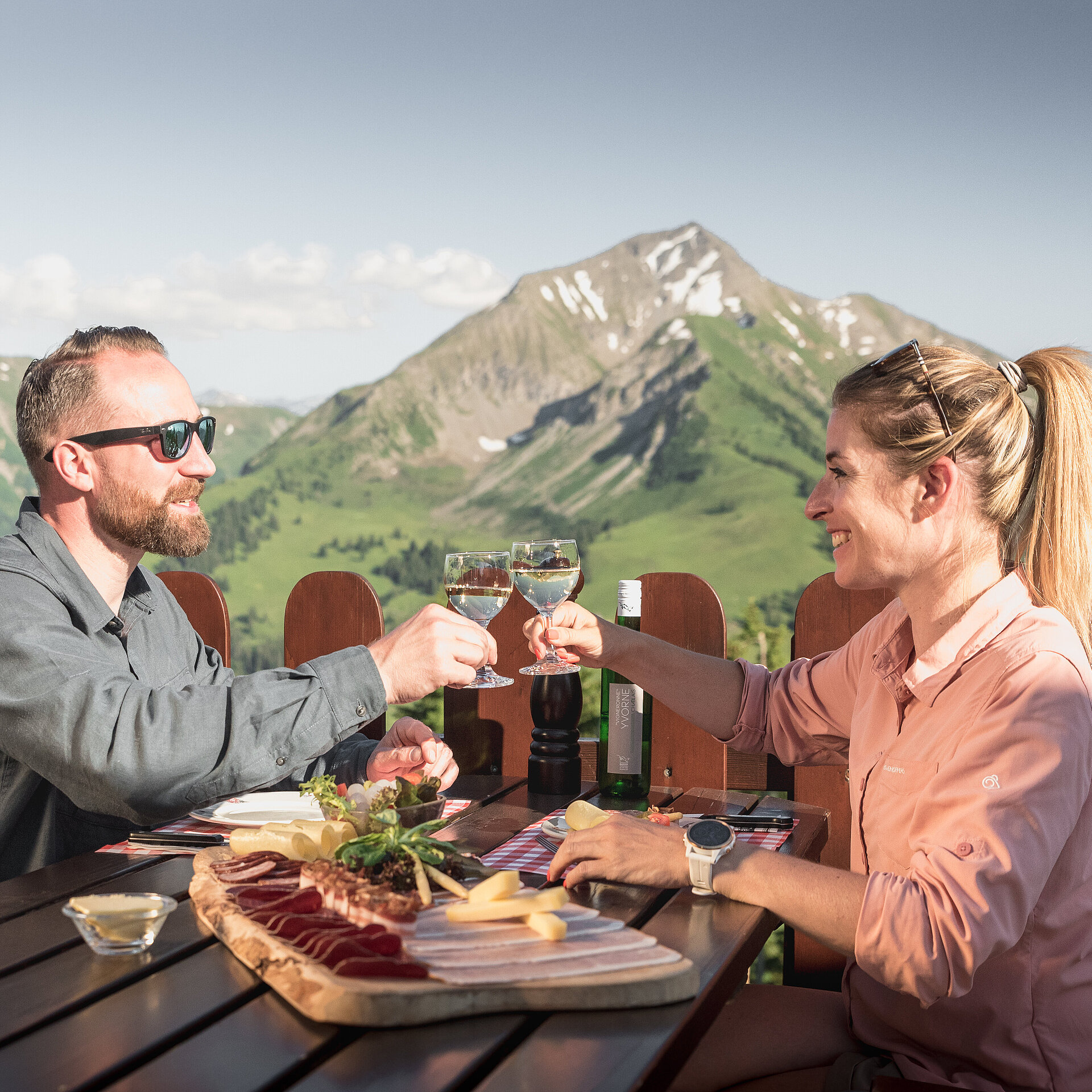 Ein paar sitzt auf einer Sonnenterrasse eines Bergrestaurants, geniesst eine Käse- und Fleischplatte und stösst mit einem Glas Weisswein an. Im Hintergrund ein Bergpanorama.