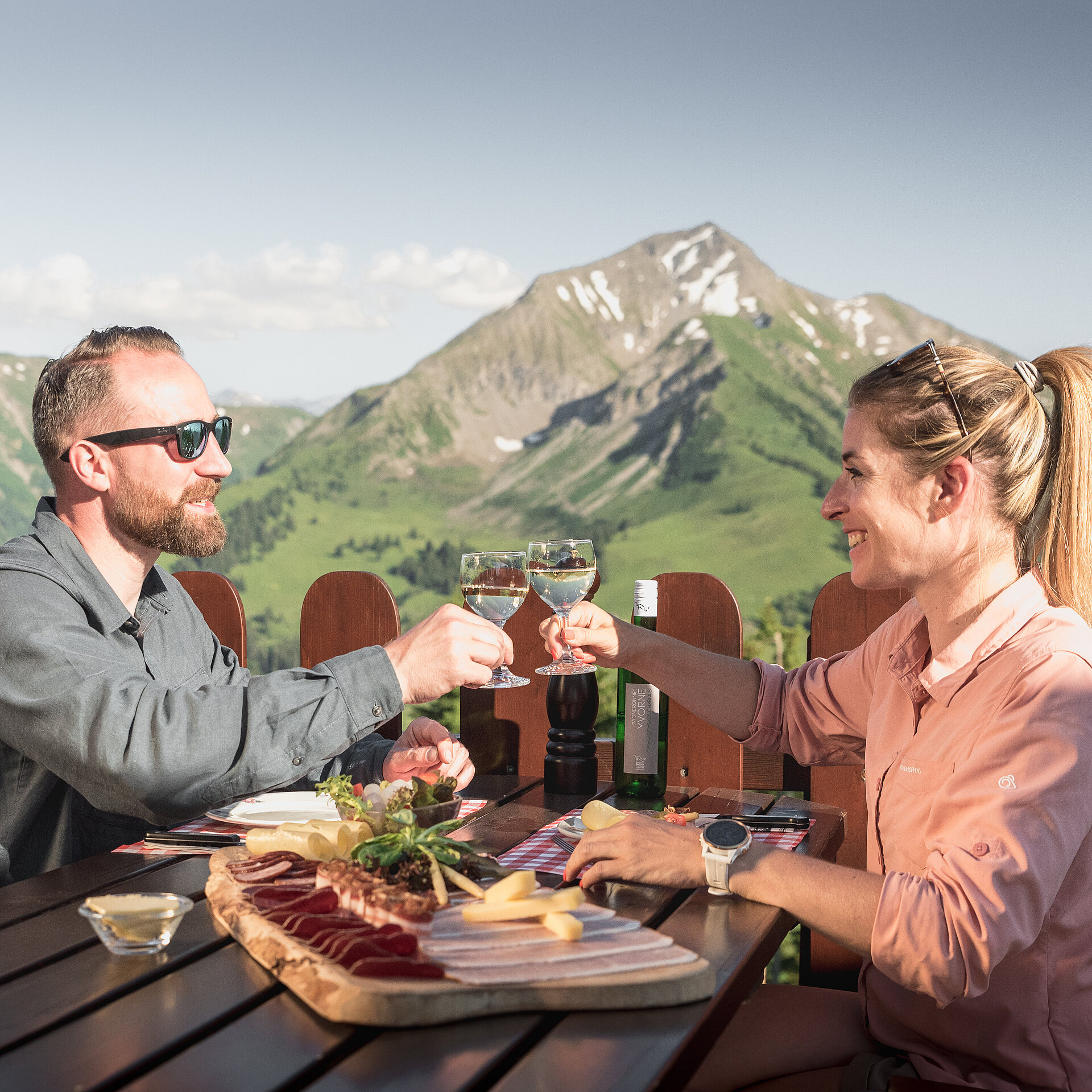 Ein paar sitzt auf einer Sonnenterrasse eines Bergrestaurants, geniesst eine Käse- und Fleischplatte und stösst mit einem Glas Weisswein an. Im Hintergrund ein Bergpanorama.