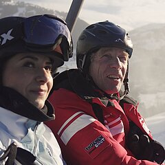 Ein Mann (Willi Bach) und eine Frau (unbekannt) sitzen mit Skiausrüstungen auf einem Sessellift und blicken beide in Richtung eines Berggipfels.