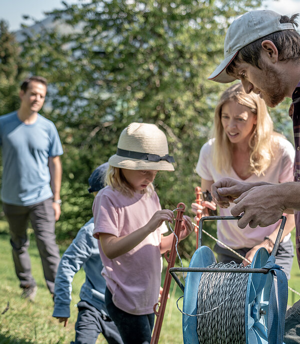 Zwei Kinder helfen einem Bauern mit dem Zaun aufstellen. Die Eltern stehen mit Gabeln etwas weiter hinten im Bild. Es ist sonnig und sie stehen auf einer grünen Wiese.