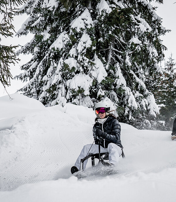 Ein Mann und eine Frau sitzen auf zwei Schlitten und fahren auf der Schlttelpiste durch den verschneiten Wald.