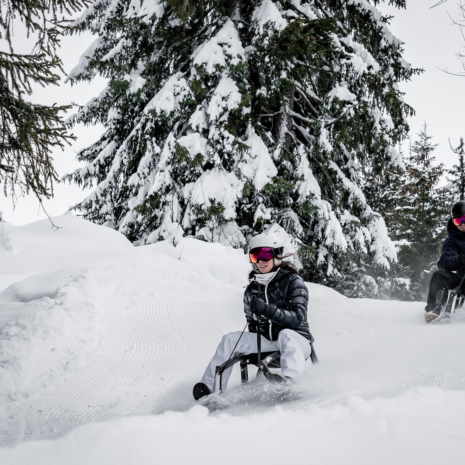 Ein Mann und eine Frau sitzen auf zwei Schlitten und fahren auf der Schlttelpiste durch den verschneiten Wald.