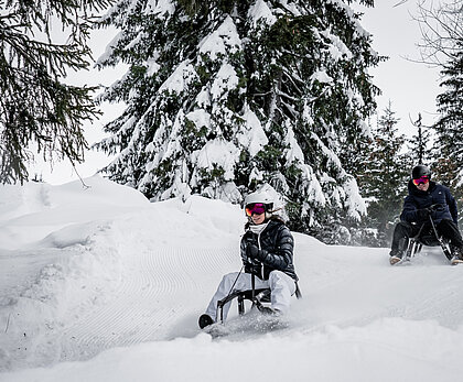 Ein Mann und eine Frau sitzen auf zwei Schlitten und fahren auf der Schlttelpiste durch den verschneiten Wald.