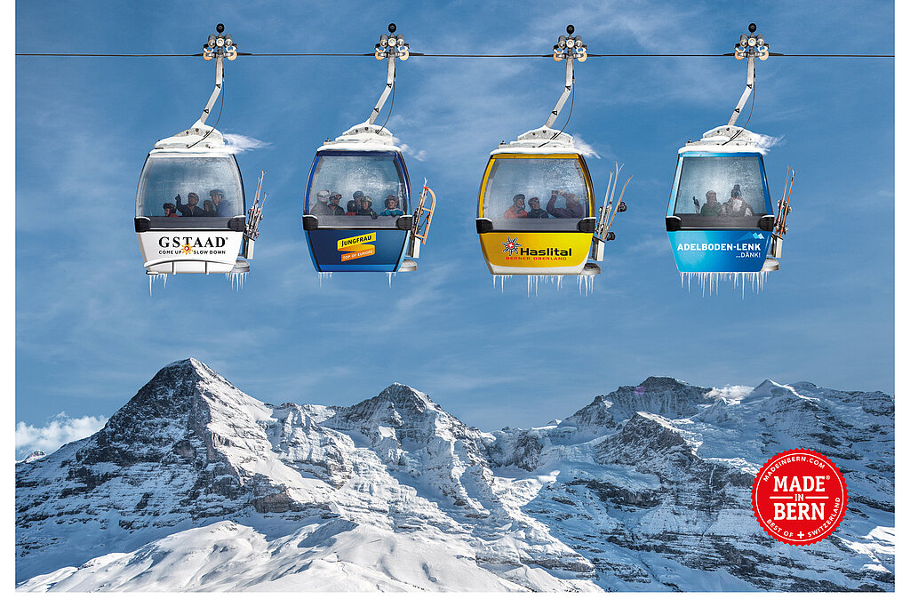 Vier Gondeln (weiss, dunkelblau, gelb und königsblau) mit den Logos der Top4-Skiregionen Gstaad, Jungfrau Ski Region, Meiringen-Hasliberg und Adelboden-Lenk ) hängen an einem Seil, darunter Bergpanorama.